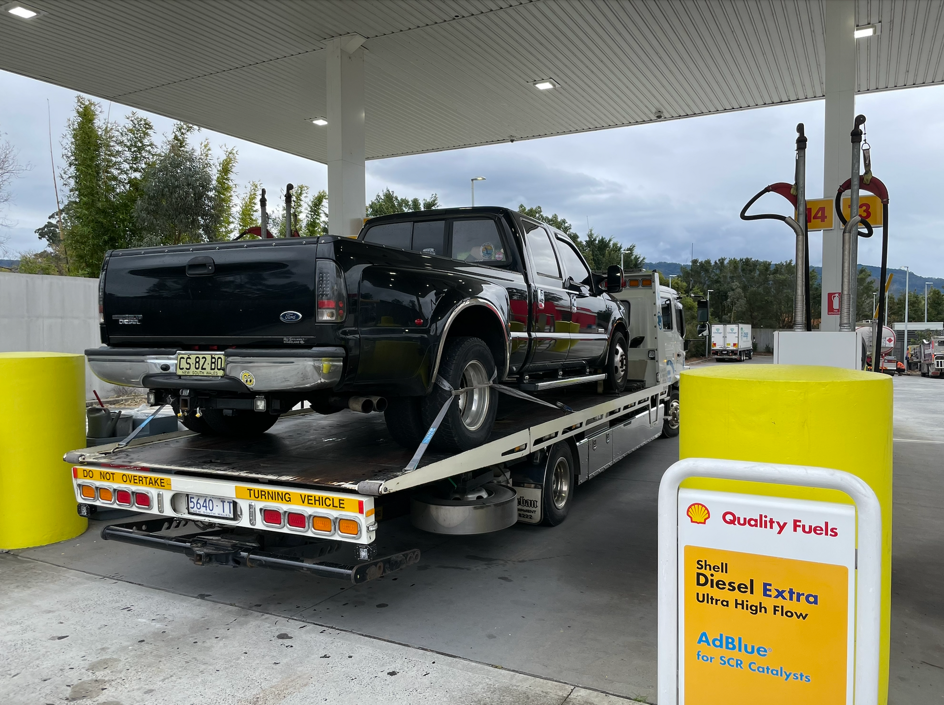 A black pickup truck being transported on a flatbed tow truck at a Shell gas station. — M.L. Wray Towing & Automotive Repairs in Nowra, NSW
