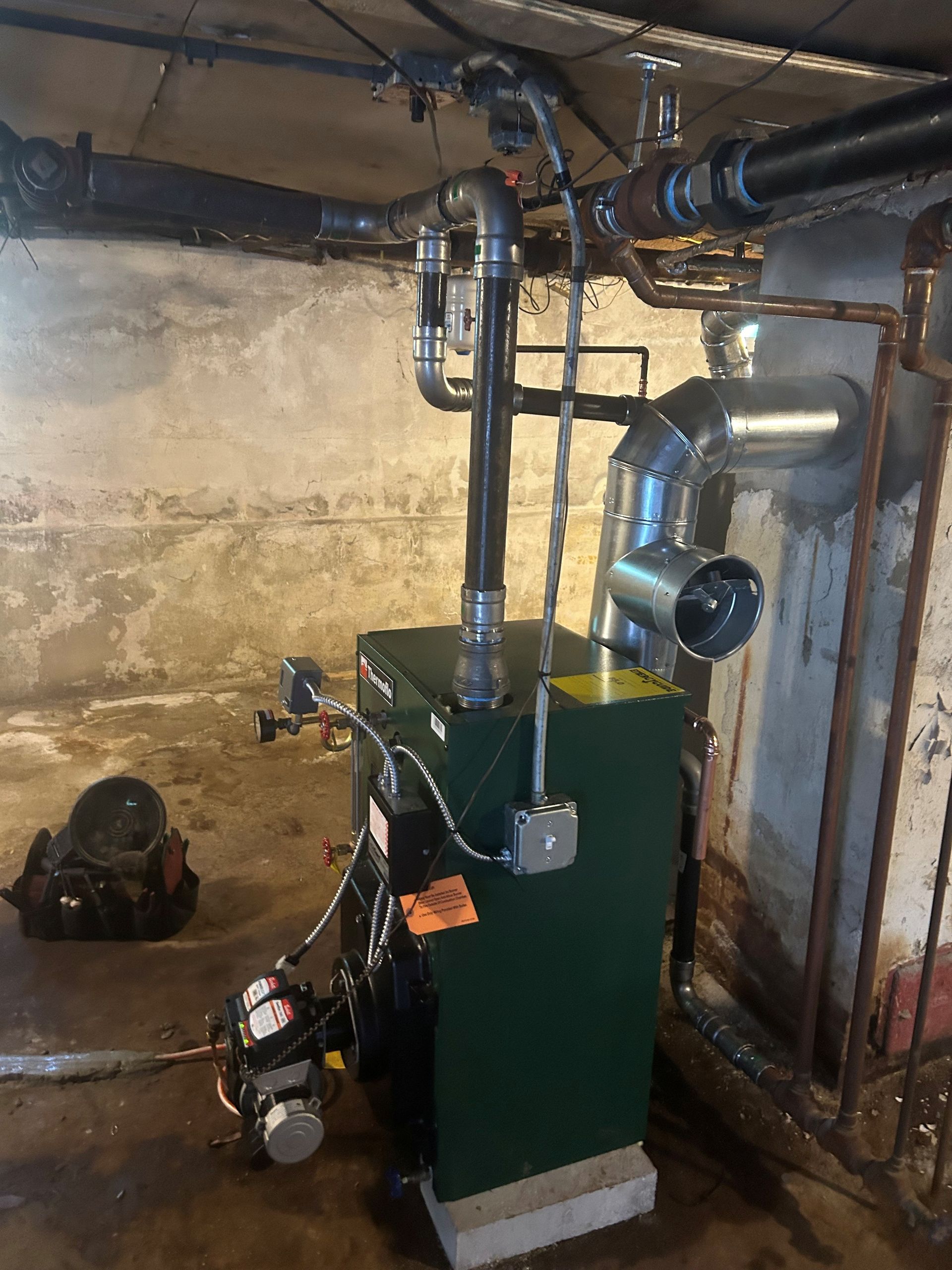 A green heating boiler in a basement, with metal pipes and wiring against a concrete wall.