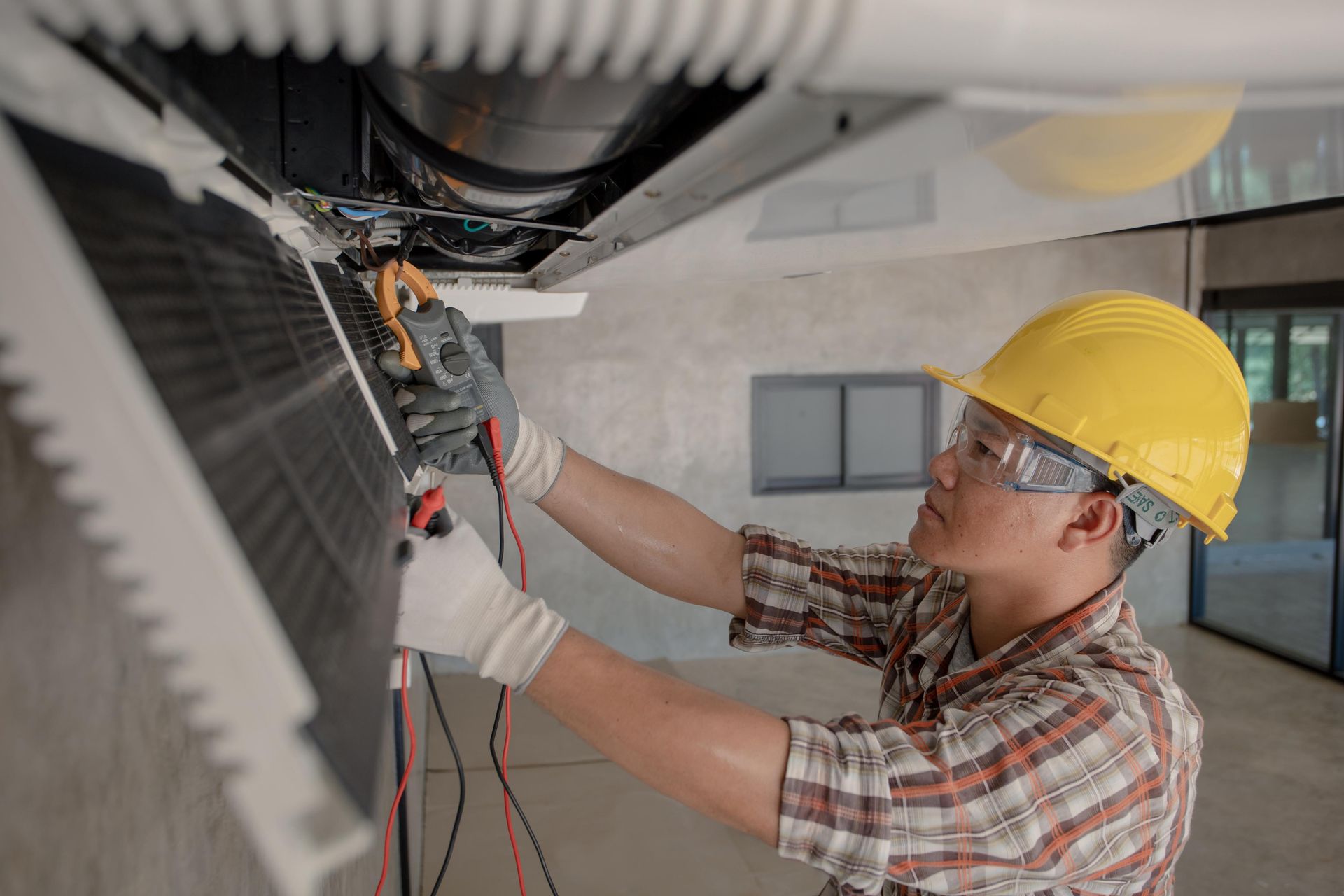 HVAC technician in yellow hard hat tests wires of ceiling unit with multimeter.