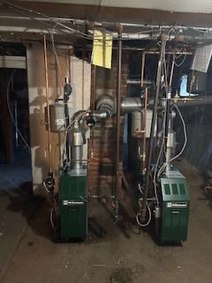 Two green boilers with copper pipes in a basement setting.