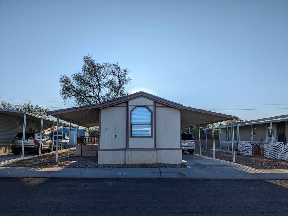 Mobile home with carports; light beige siding, blue window, cars parked in front.