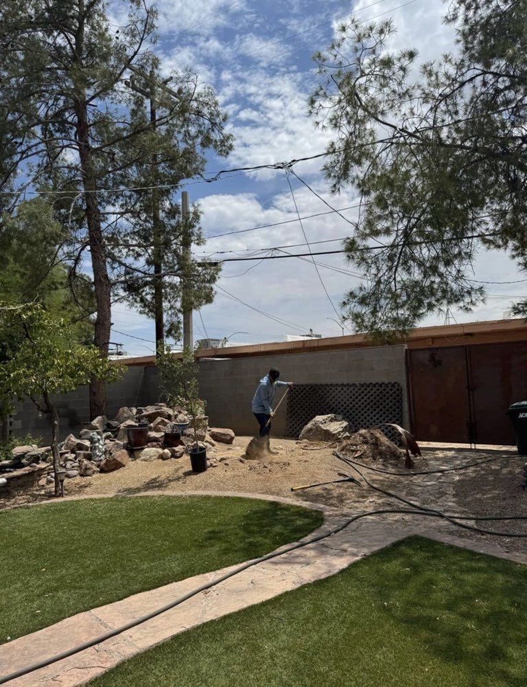 Person shoveling dirt in a backyard with trees, a wall, and a patch of grass on a sunny day.