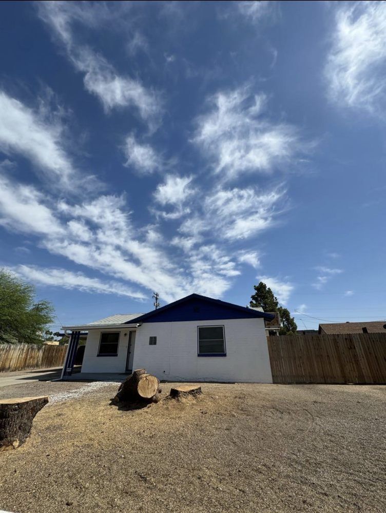 White house with blue roof under a blue sky with clouds, surrounded by a fence and gravel.