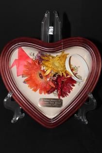 Heart-shaped red-framed display with pressed flowers and a small plaque on a clear stand
