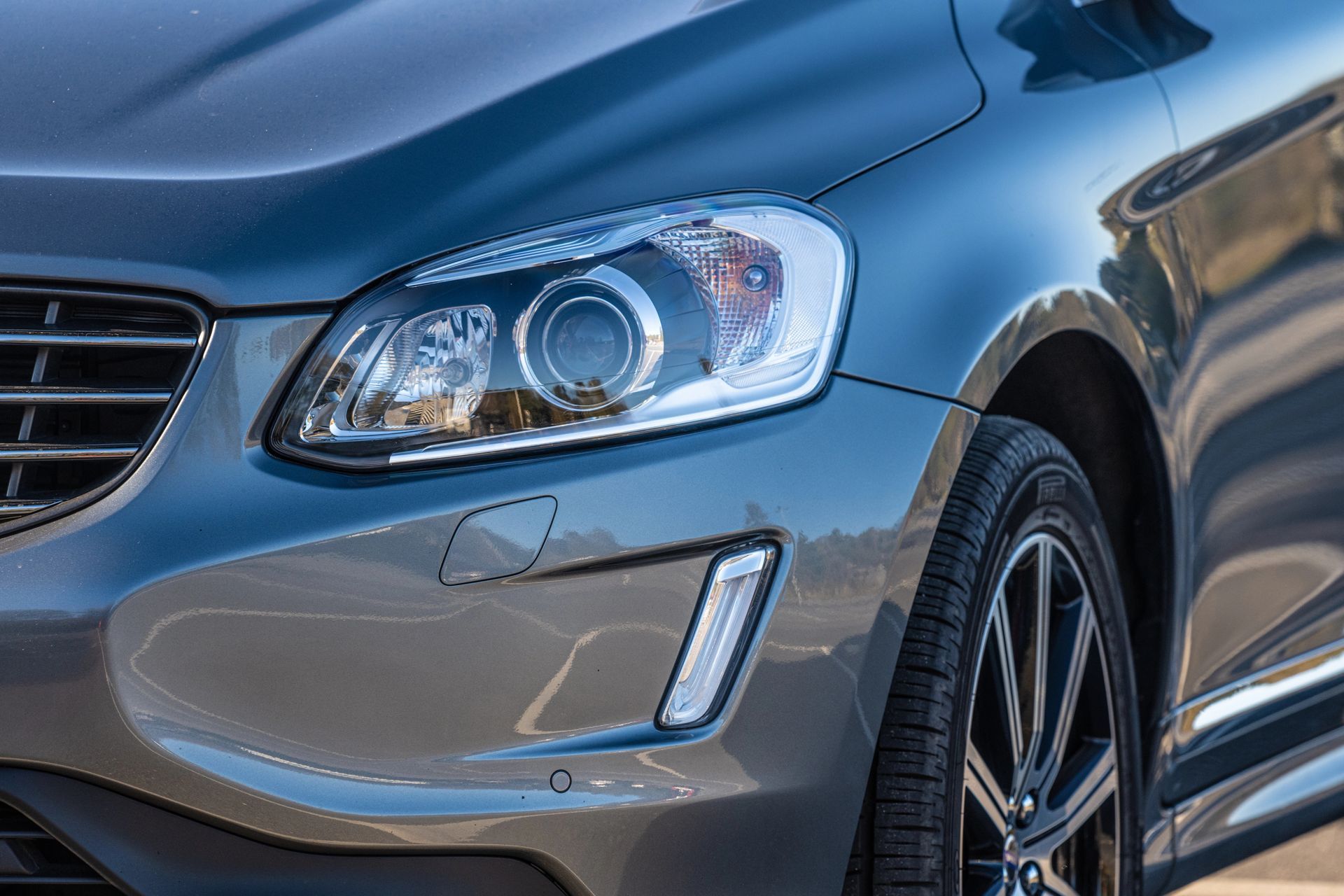 Primo piano del faro anteriore lato passeggero, della griglia e del paraurti di una Volvo XC60 grigio scuro.