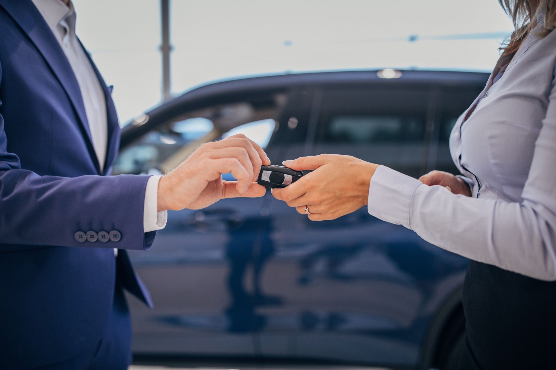 In una concessionaria, con un'auto sullo sfondo, una persona in abito da lavoro consegna le chiavi di un'auto a un'altra.