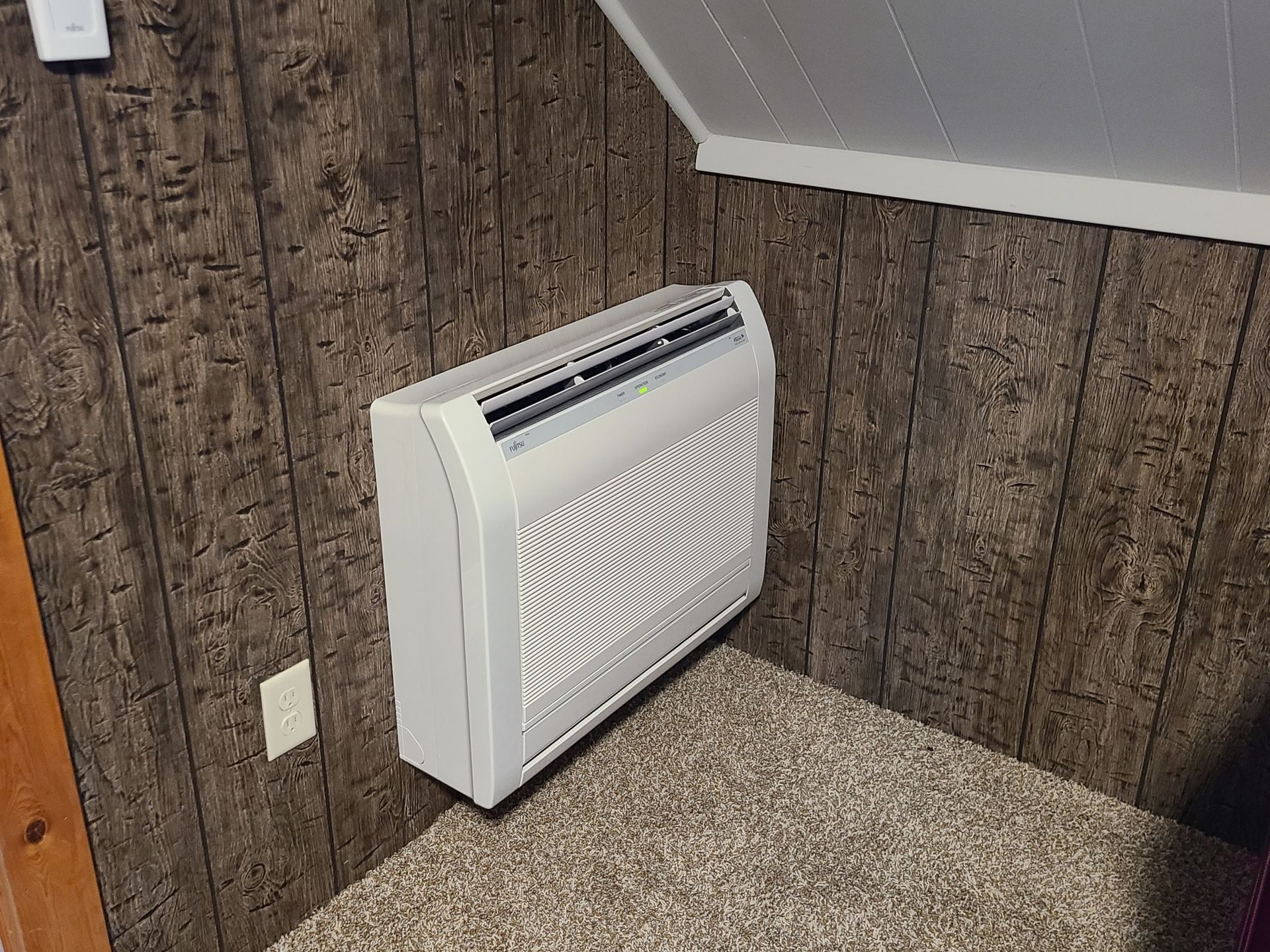 A white air conditioner is sitting on a carpeted floor in a room.