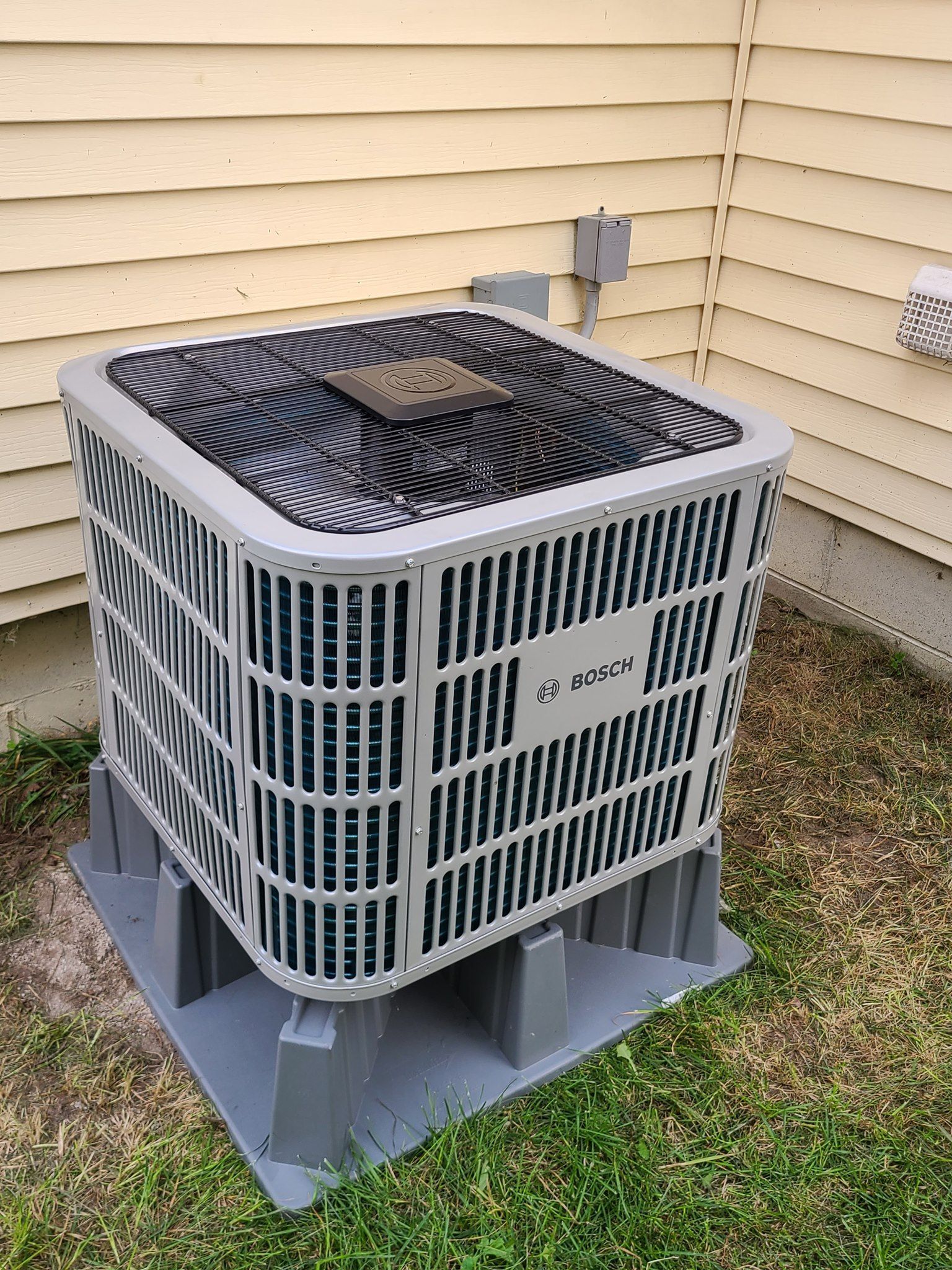 A gray air conditioner is sitting in the grass next to a house.