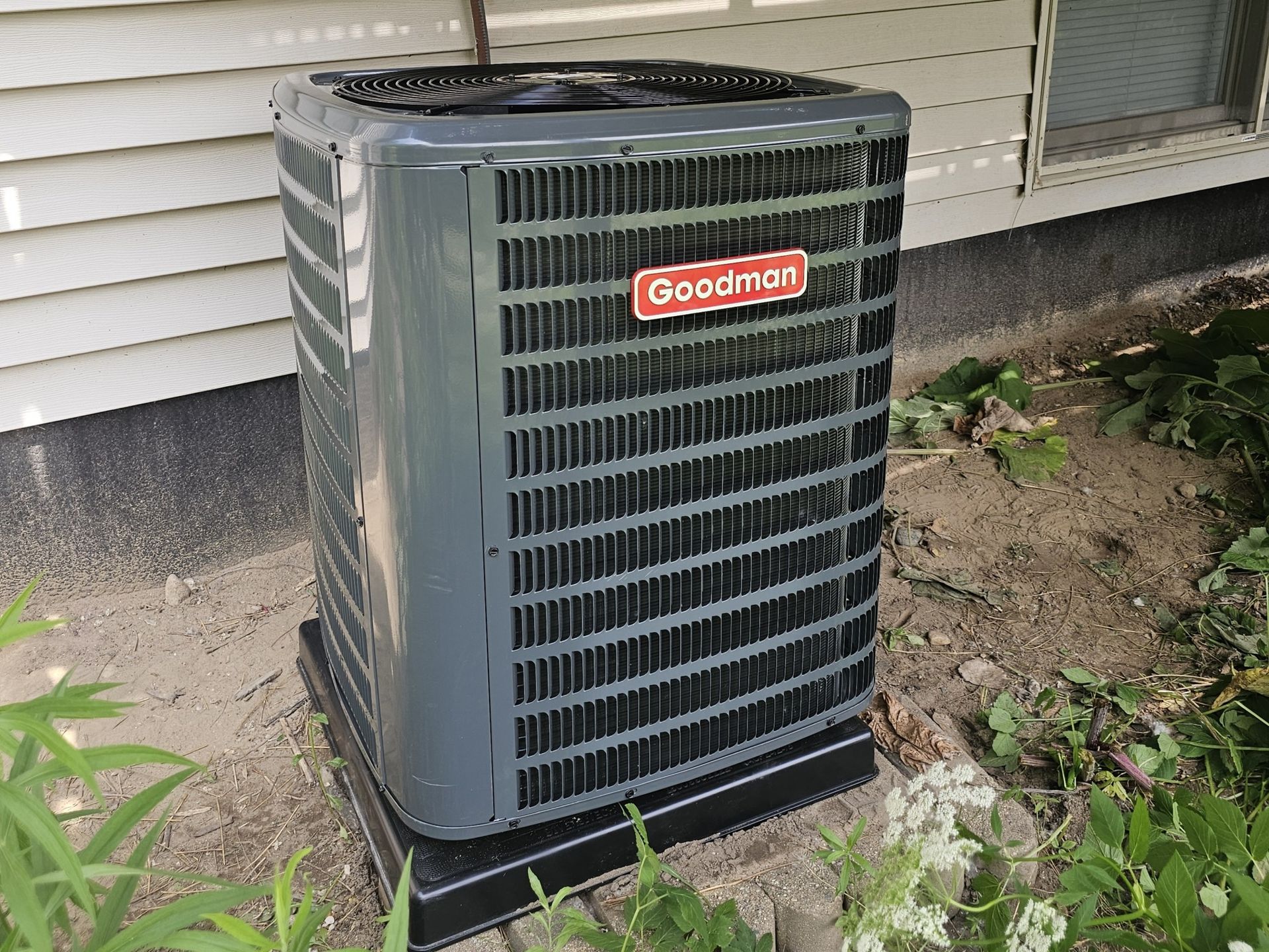 A goodman air conditioner is sitting outside of a house.