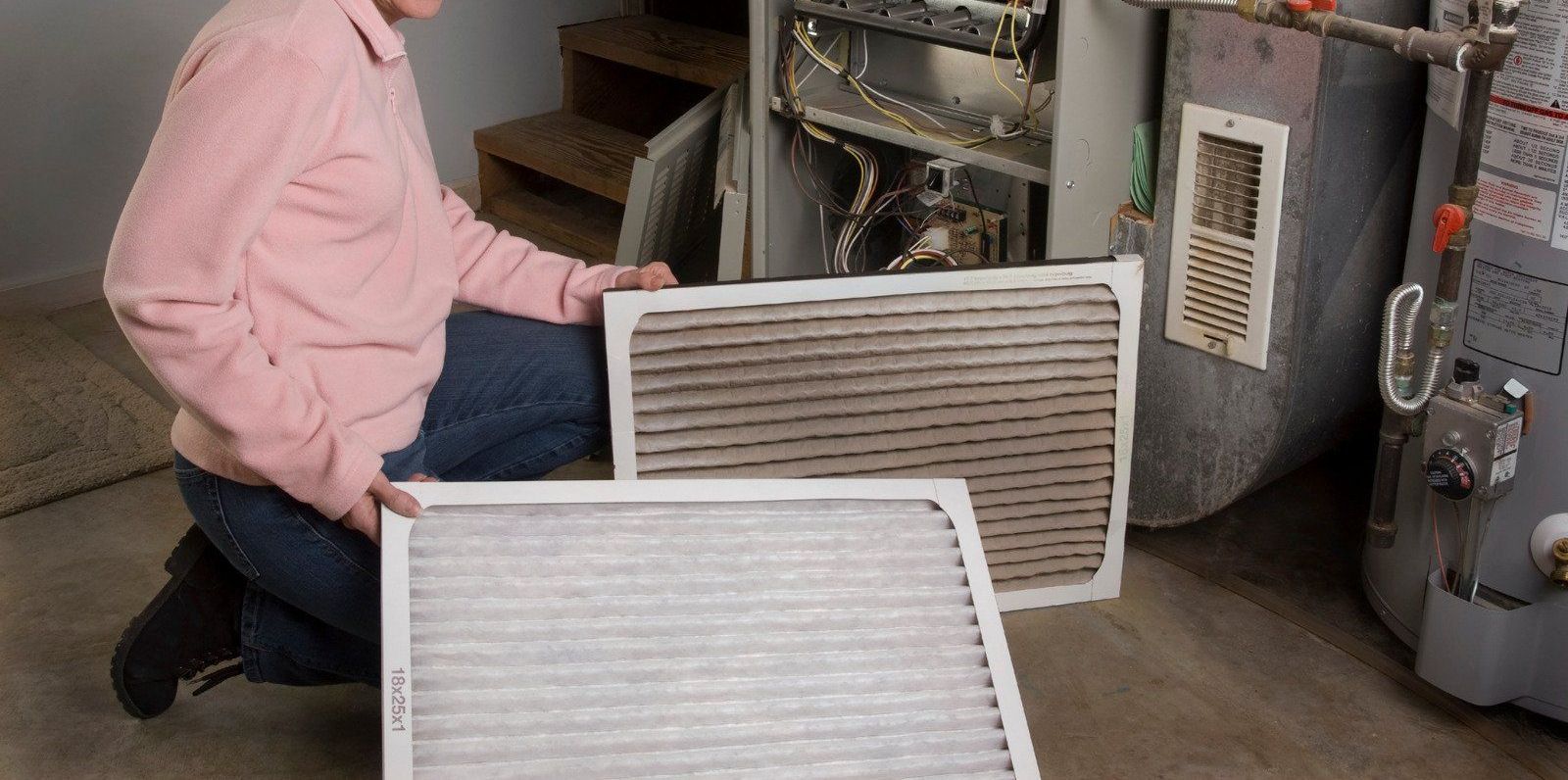 A man is kneeling down in front of a furnace holding two filters.