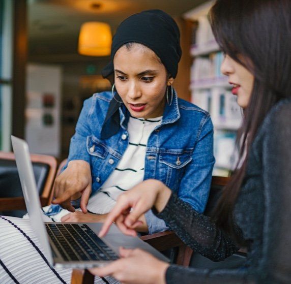 Two women looking at a laptop, pointing. One wears a denim jacket and head covering. Indoors, cafe setting.