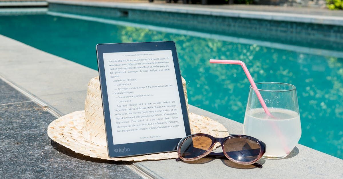 A tablet , sunglasses , a hat and a glass of water are sitting next to a swimming pool.