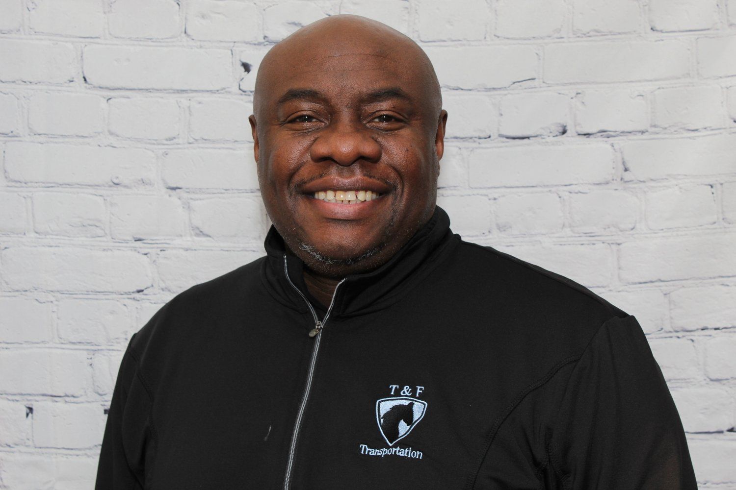 A man in a black jacket is smiling in front of a white brick wall.
