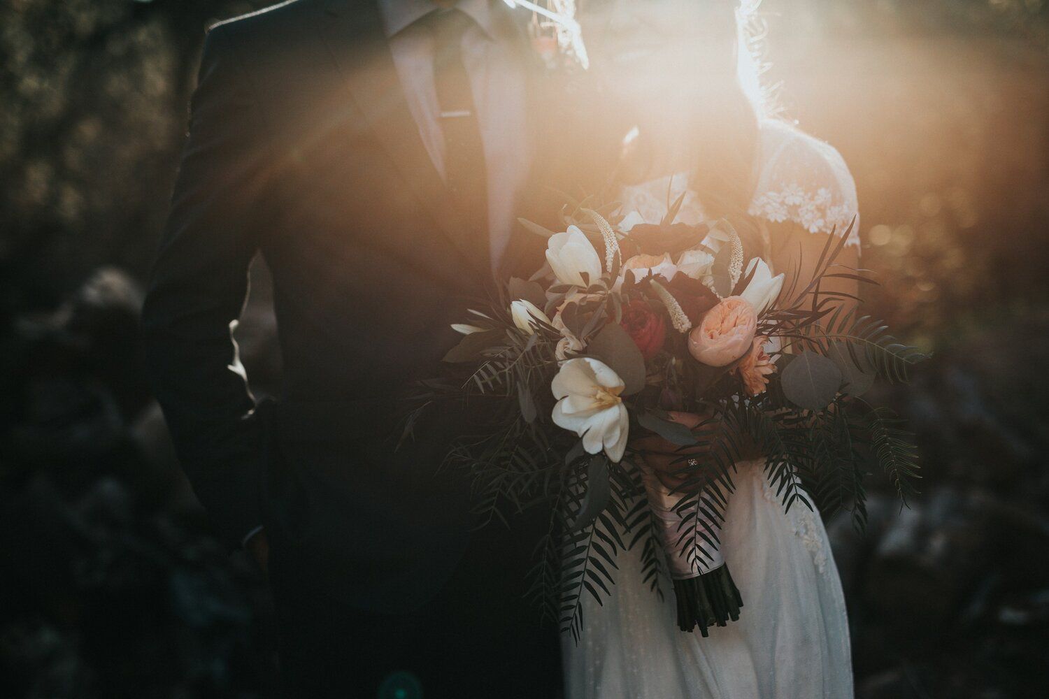 A bride and groom are standing next to each other in a field holding hands and a bouquet of flowers.