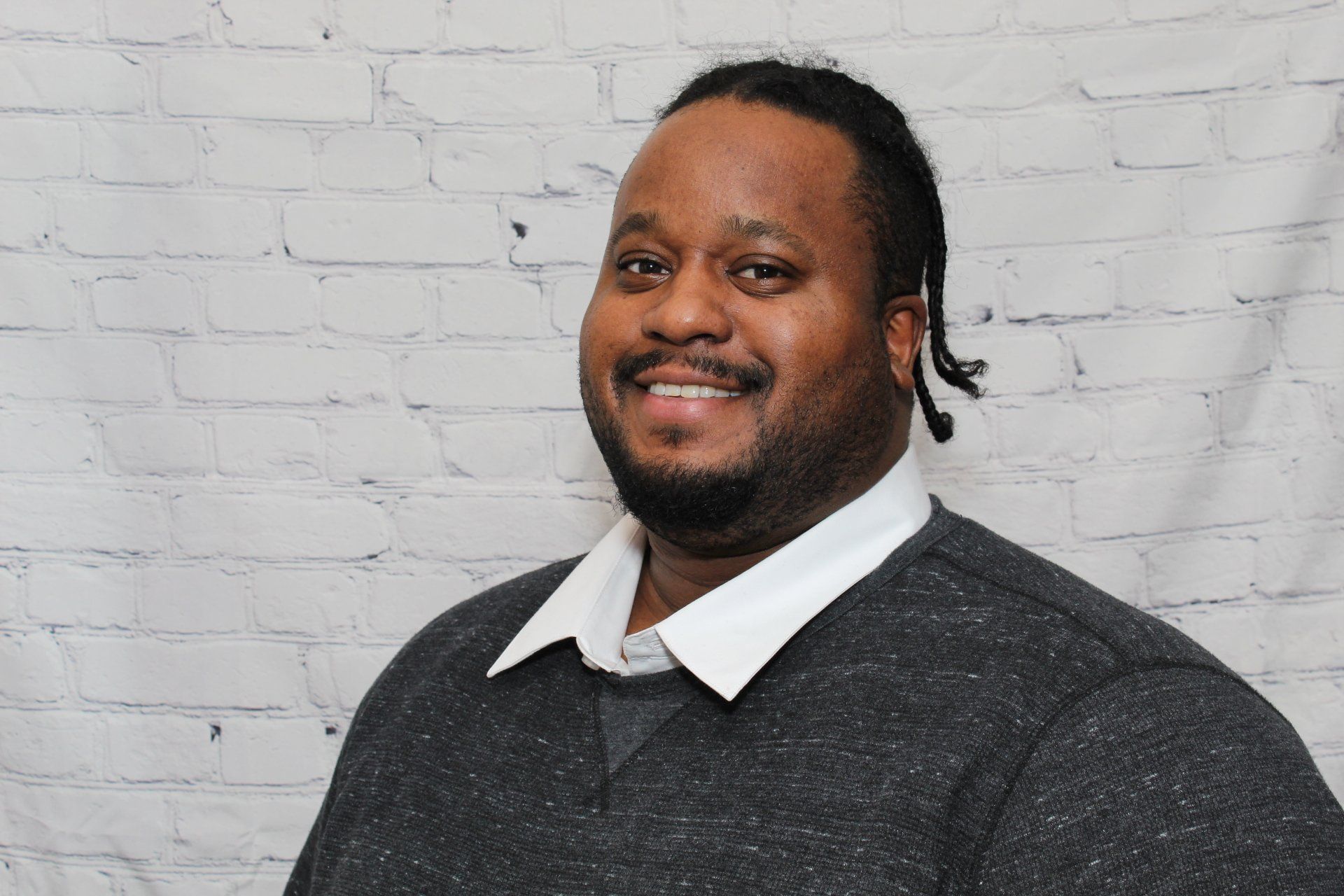 A man in a grey sweater and white shirt is smiling in front of a white brick wall.