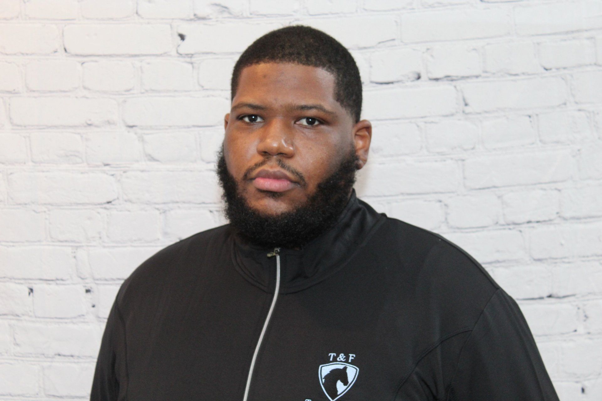 A man with a beard is standing in front of a white brick wall.