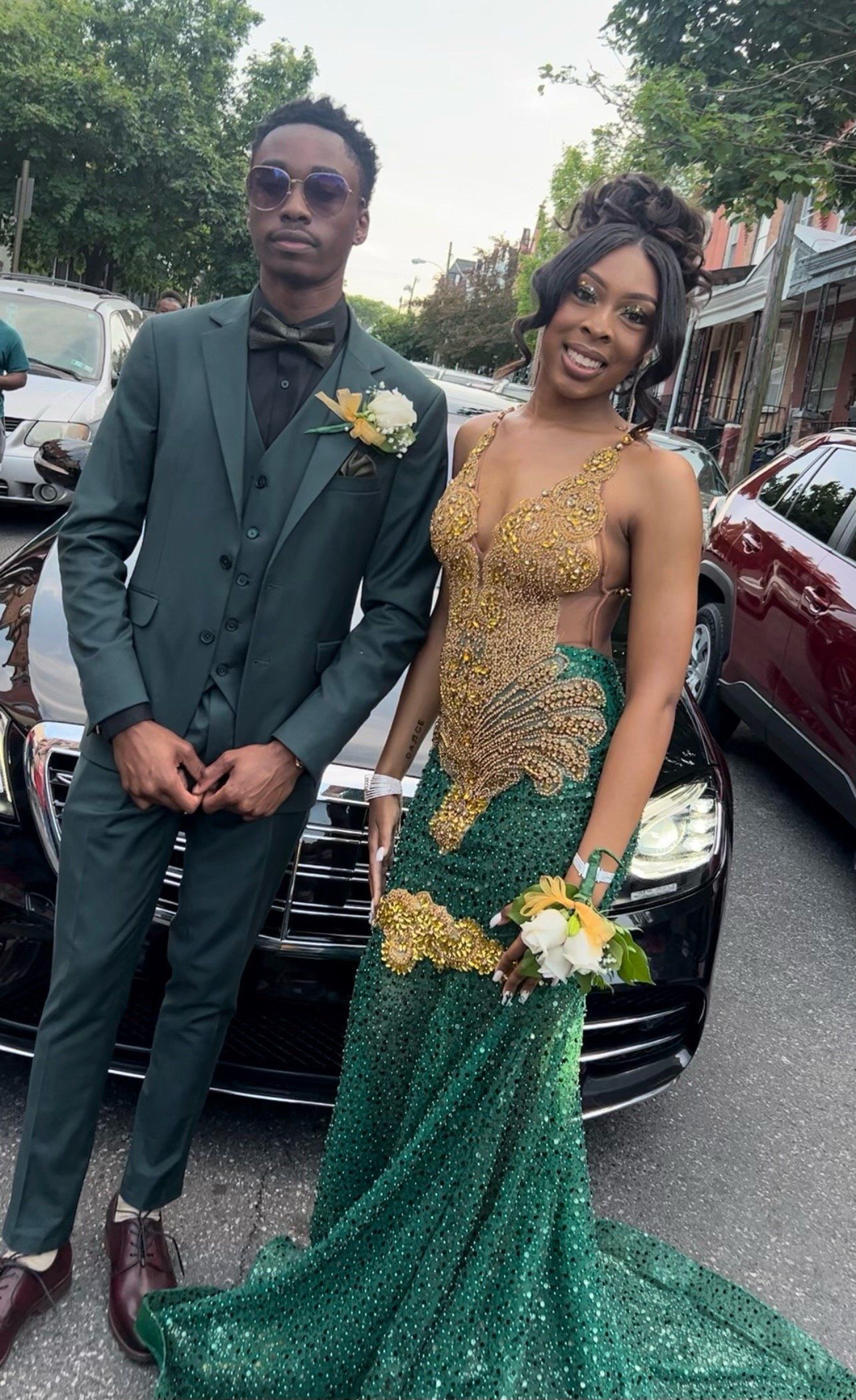 A man in a suit and a woman in a green dress are standing next to each other in front of a car.