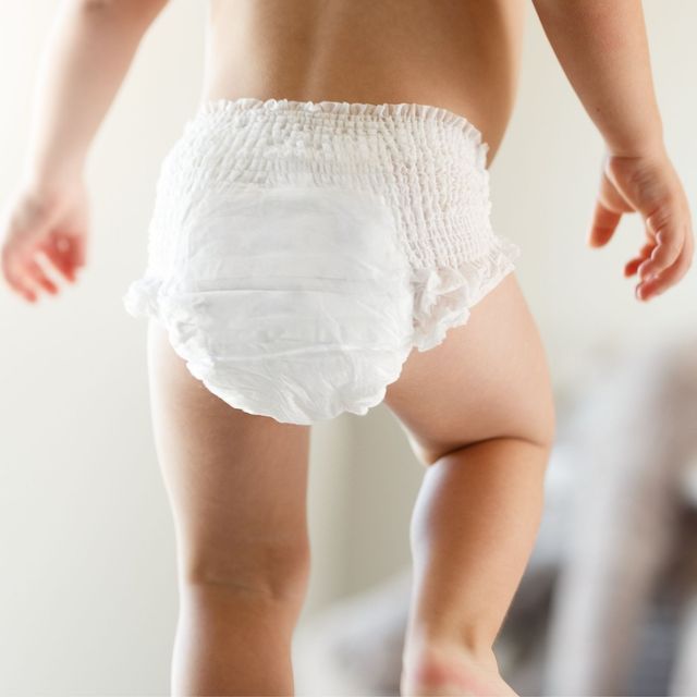 Toddler in a white diaper standing indoors, seen from behind with one leg lifted.