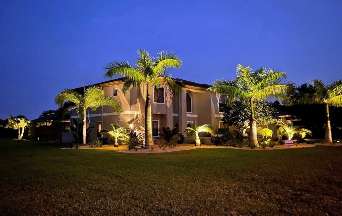A large house is lit up at night with palm trees in front of it.
