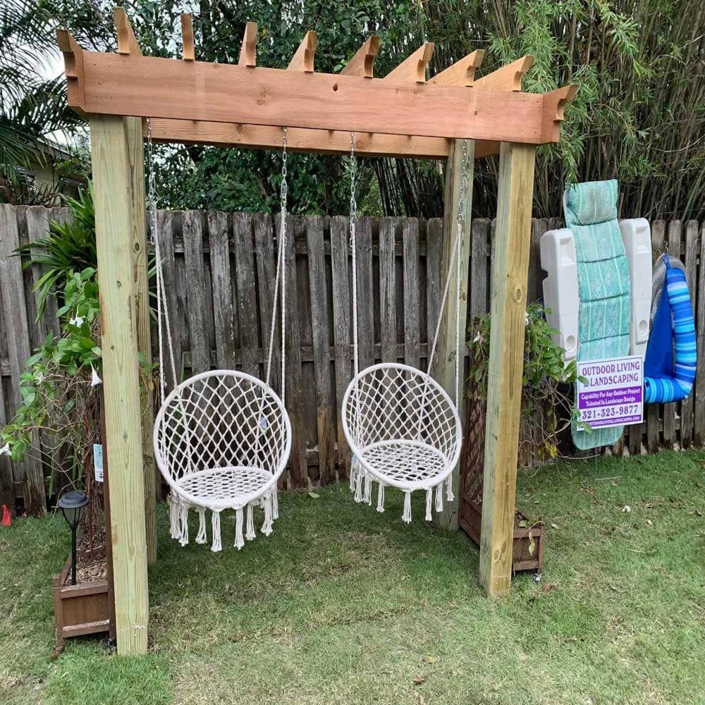 Two hanging chairs are sitting under a wooden pergola in a backyard.