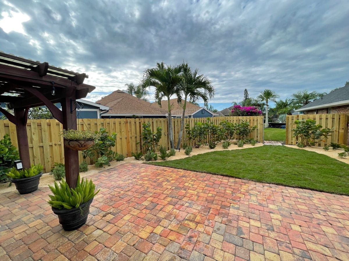 A backyard with a wooden fence , brick walkway , and potted plants.