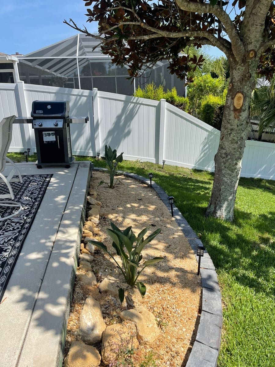A backyard with a white fence , a grill and a tree.