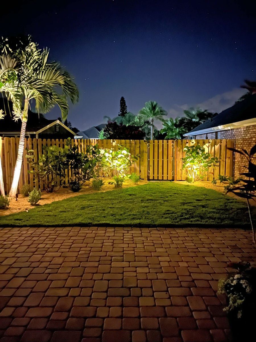 A backyard with a wooden fence and a brick patio at night.