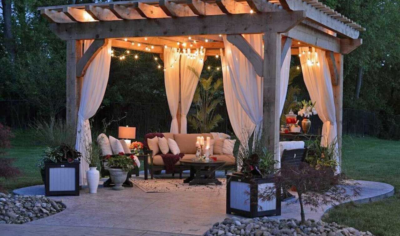 Romantic, wooden pergola with white curtains, string lights, and seating area on a stone patio.