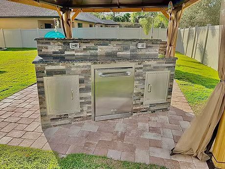 A large outdoor kitchen with a stainless steel dishwasher under a gazebo in a backyard.