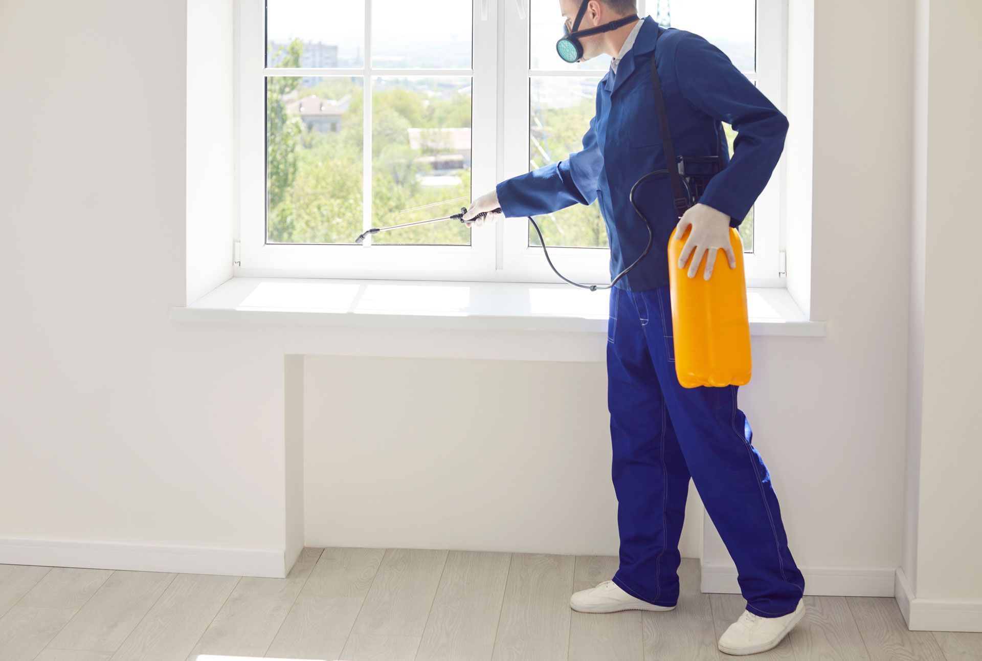 Pest control worker in blue coveralls, mask, and gloves sprays insecticide near a window in a room.