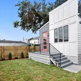 A modern white house with a red door, a gray deck with black railings, and a wooden fence bordering a grassy yard.