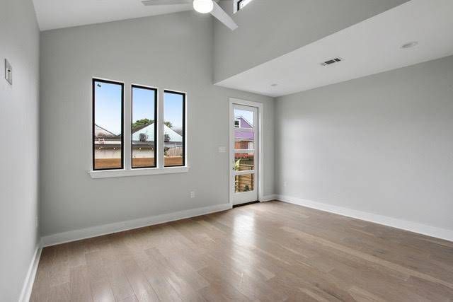 A modern, empty room with light gray walls, hardwood floors, a vaulted ceiling, a ceiling fan, and a glass-paned door.