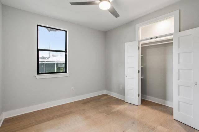 An empty room with light gray walls, light hardwood floors, a window, a ceiling fan, and an open white closet.