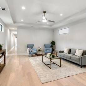 Modern living room with light wood floors, a gray sofa, two blue armchairs, a glass coffee table, and a ceiling fan.