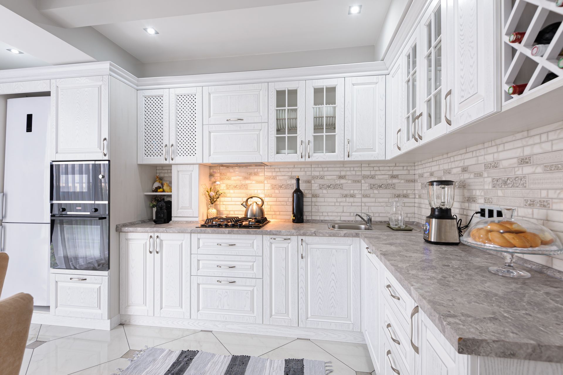A bright, white kitchen with custom cabinets, a built-in oven, stone-textured countertops, and a subway tile backsplash.