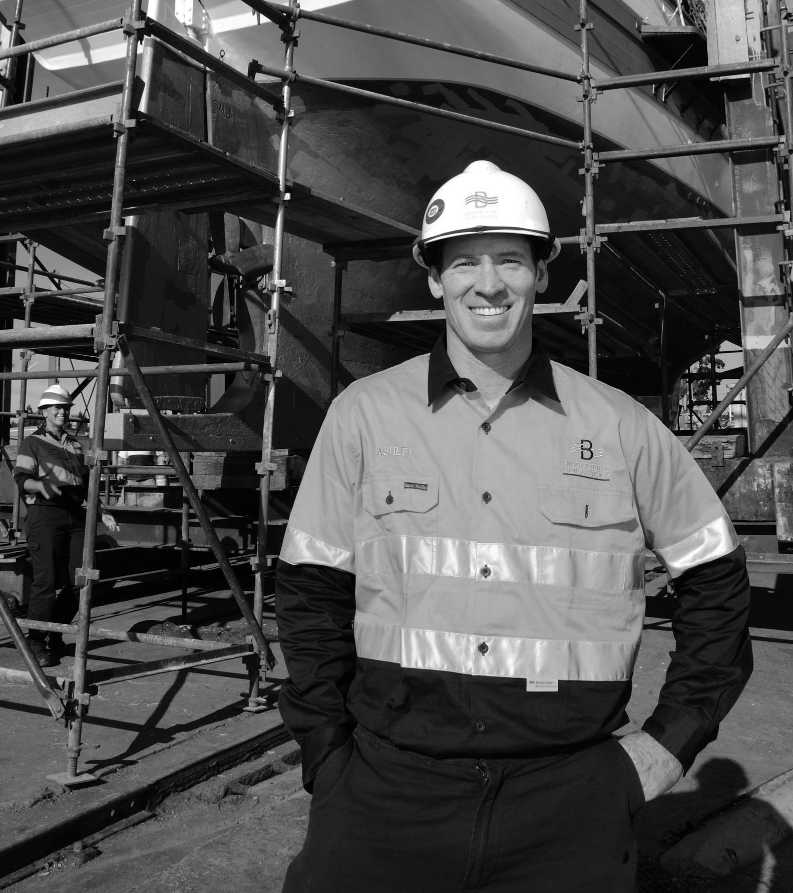 Man in work uniform smiles at camera, in front of scaffolding and ship.