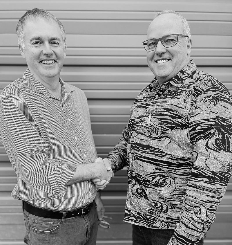 Two men shaking hands, smiling. One in striped shirt, the other in patterned shirt, in front of a wall.