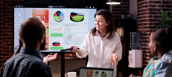 Woman presenting data to colleagues in a modern office with charts on a large screen.