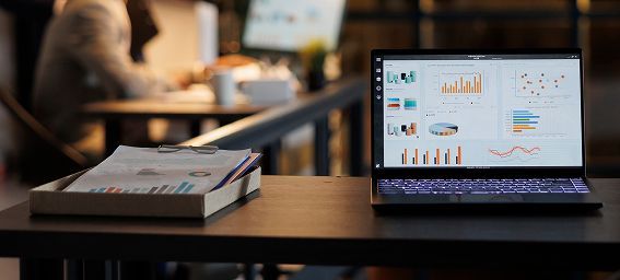 Laptop displaying data charts, papers in tray on desk, another person blurred in the background, office setting.