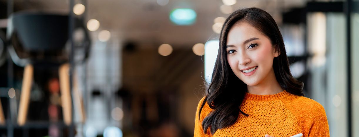 Young woman with dark hair smiles, wearing an orange sweater in an office setting.