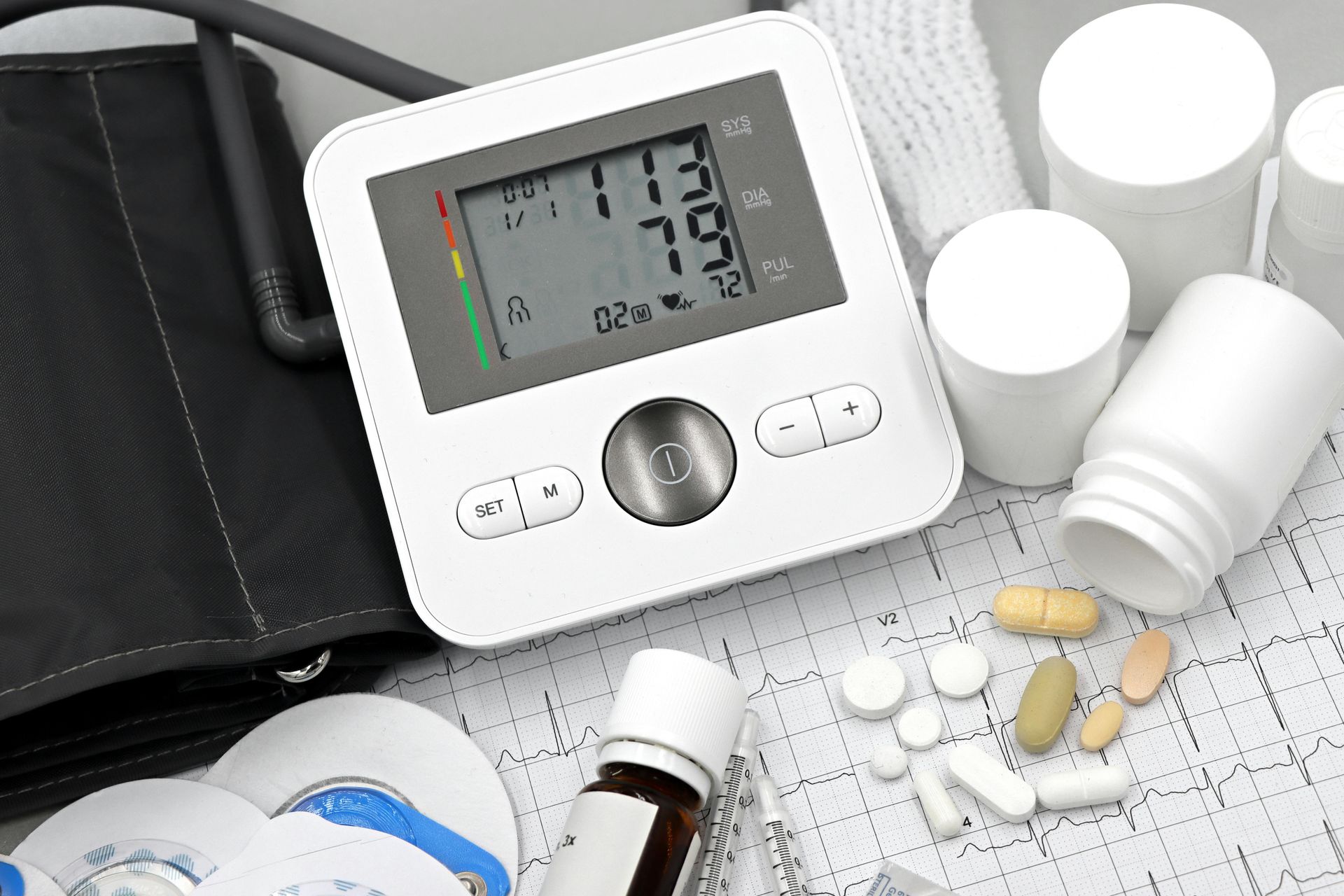 Blood pressure monitor displaying readings alongside various medication bottles and pills on an ECG paper, representing remote patient monitoring for chronic disease management.
