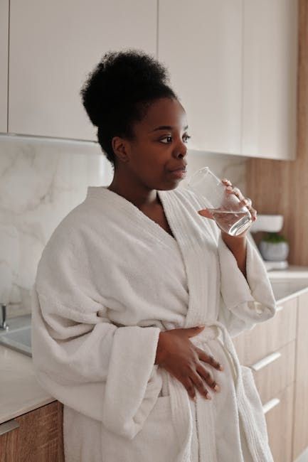 Woman in a white bathrobe drinking water in a kitchen, emphasizing hydration for managing constipation related to GLP-1 medications.