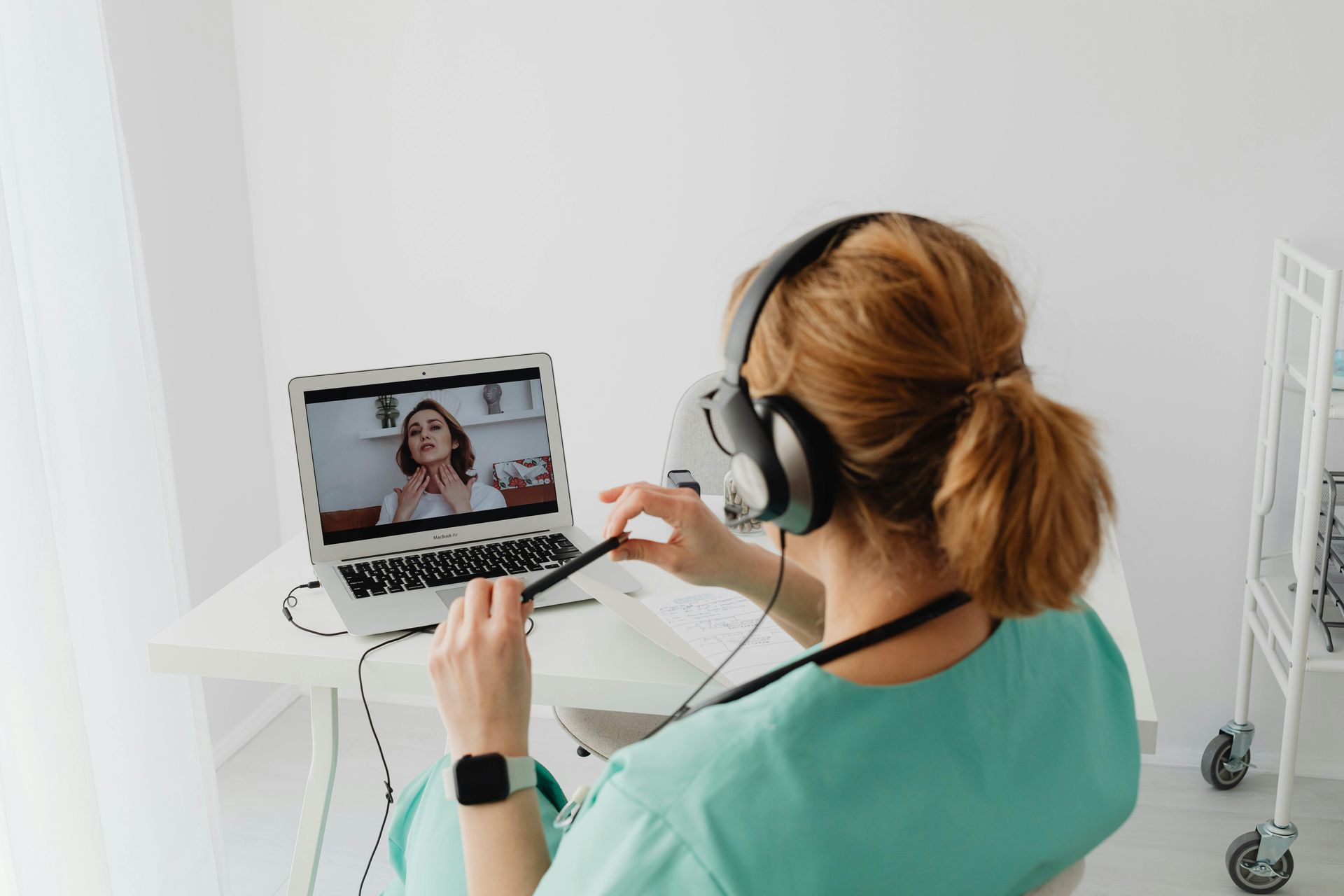 Healthcare professional in teal scrubs using a laptop for telehealth consultation, engaging with a patient discussing skin condition.