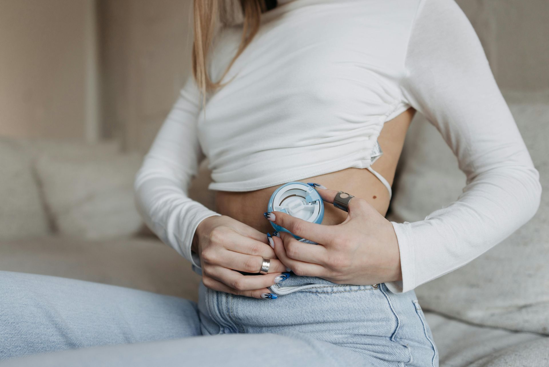 Woman wearing a white long-sleeve top applying a medical device to her abdomen, illustrating the use of advanced wearable technology for health monitoring.