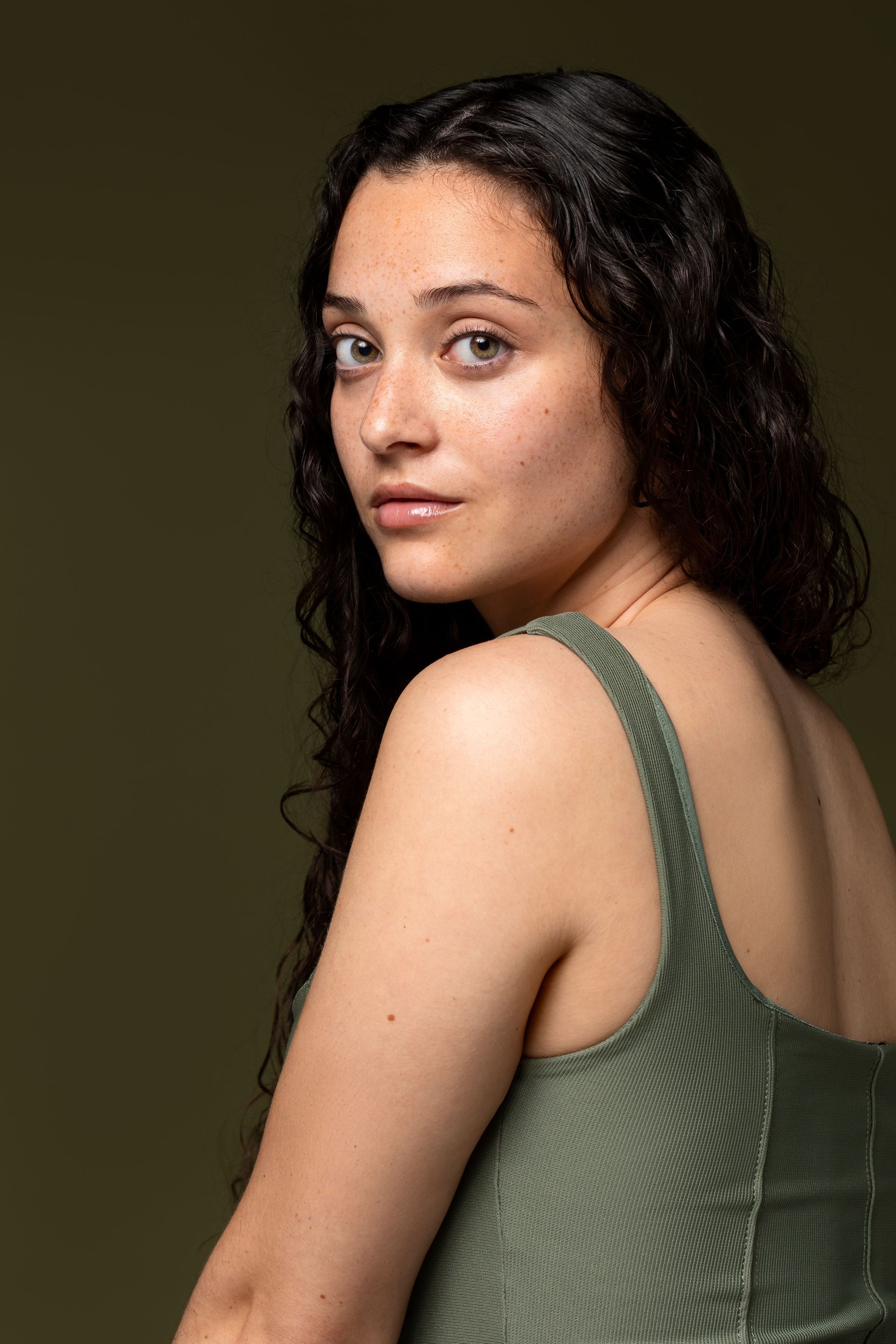 Woman with curly hair, looking over her shoulder. Wearing a green dress against a green background.