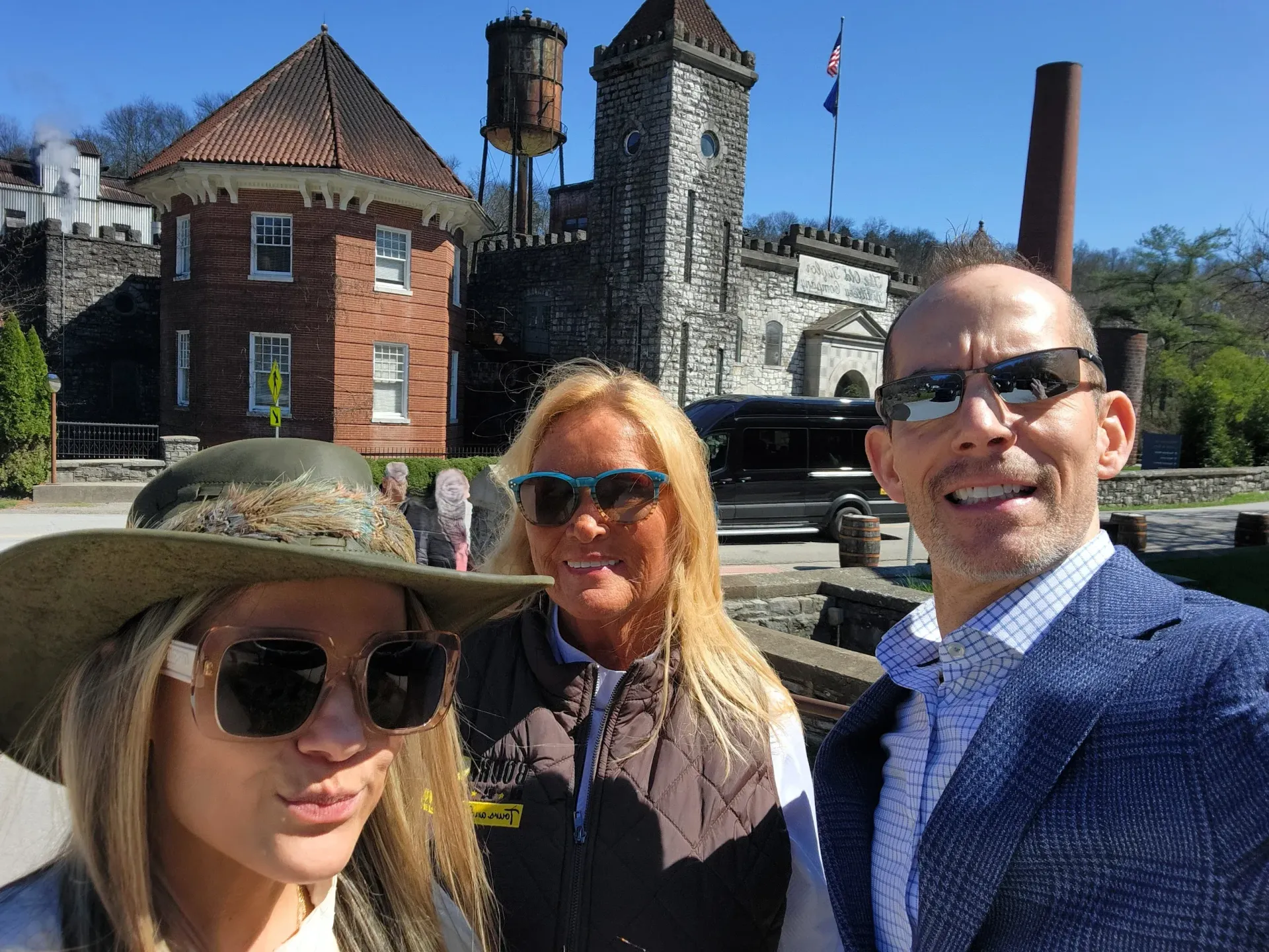 Three people posing outdoors in front of a brick building and stone tower under a blue sky.