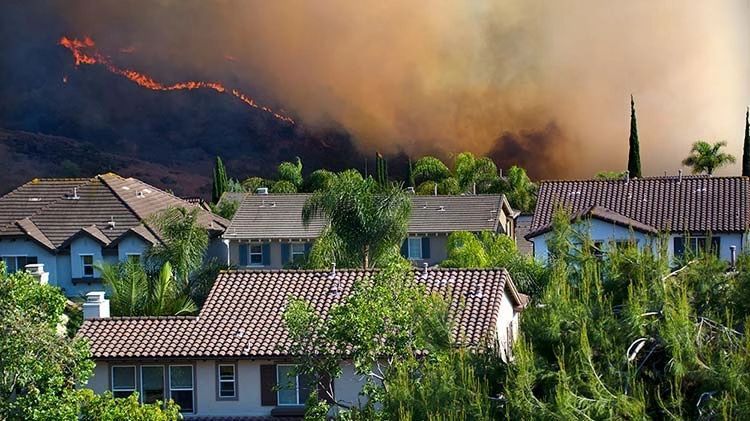 Houses in foreground with a wildfire burning on a hillside in the background, smoke filling the sky.