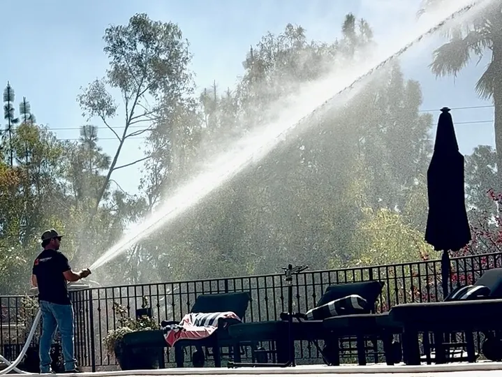 Man spraying water with a hose on a sunny day near a pool, with trees in the background.