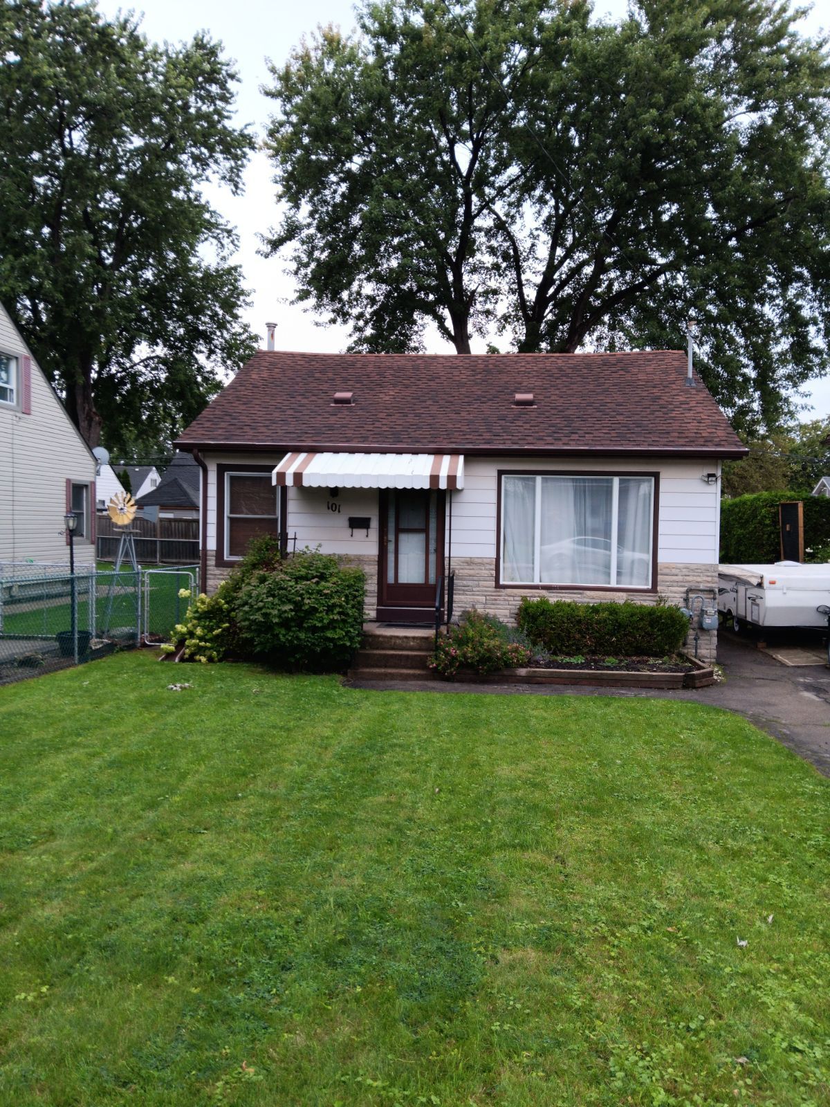 Small, white house with a brown roof, small awning, and green lawn, trees in background.