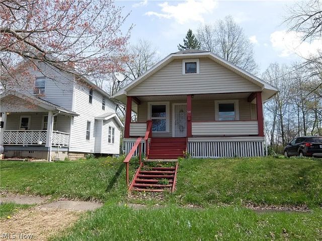 Tan house with red porch and steps, next to a white house, on a grassy hill.